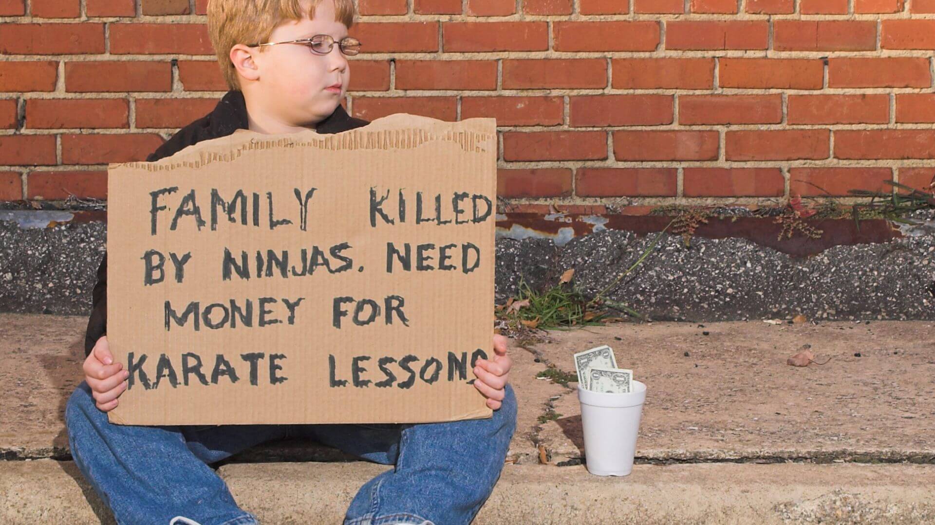 A boy holding a hand-written sign "Family killed by ninjas. Need money for karate lessons"