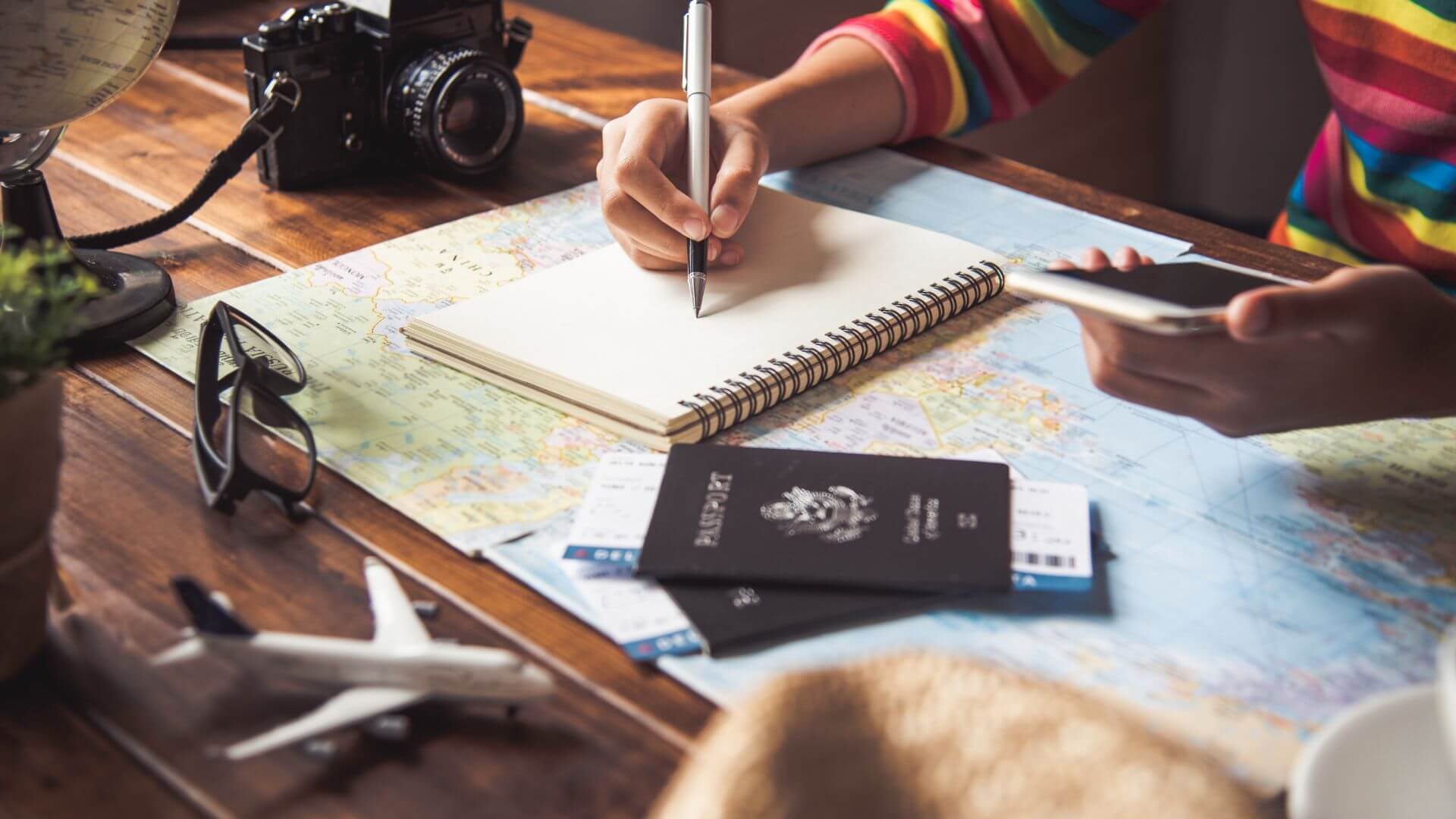 A person sitting at the table planning a vacation. There is a map, a passport, and two plane tickets on the table. This person is writing something dowin into an empty notebook.