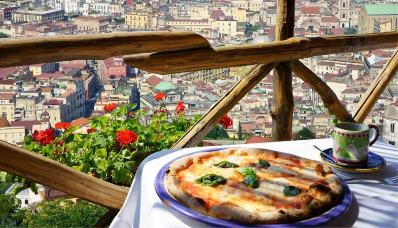 A photo if pizza on a balcony with a view over Naples, a must-visit city during Italy tour